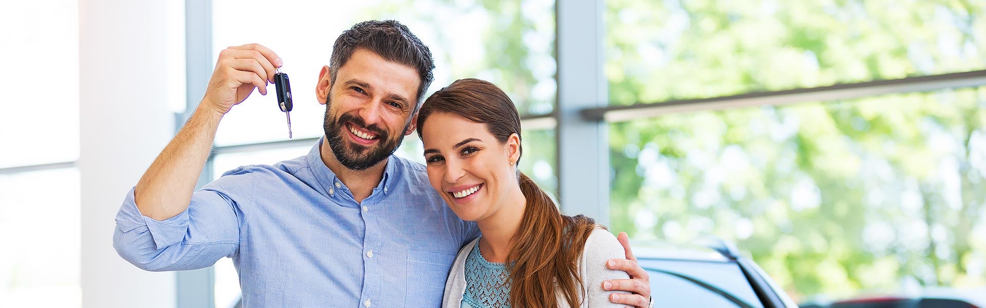 A happy couple in a car dealership, the man holding up new car keys, smiling at the camera.